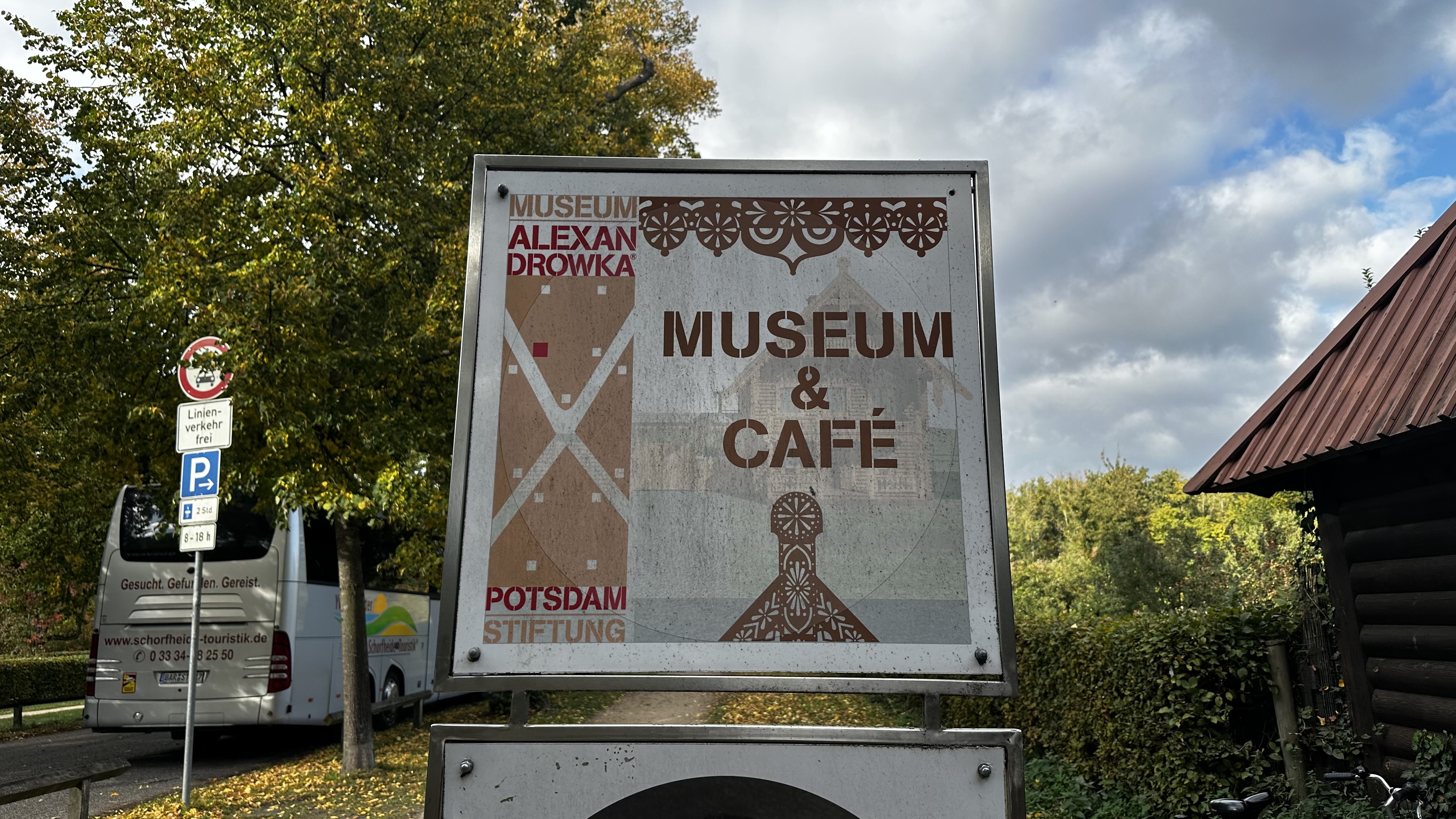 Ein Schild mit der Aufschrift „Museum & Café“ und dekorativen Mustern im Freien, umgeben von Bäumen und einem geparkten Bus – ein einladender Zwischenstopp während Ihres Ausflugs nach Potsdam, um die lokalen Sehenswürdigkeiten der Stadt zu entdecken.