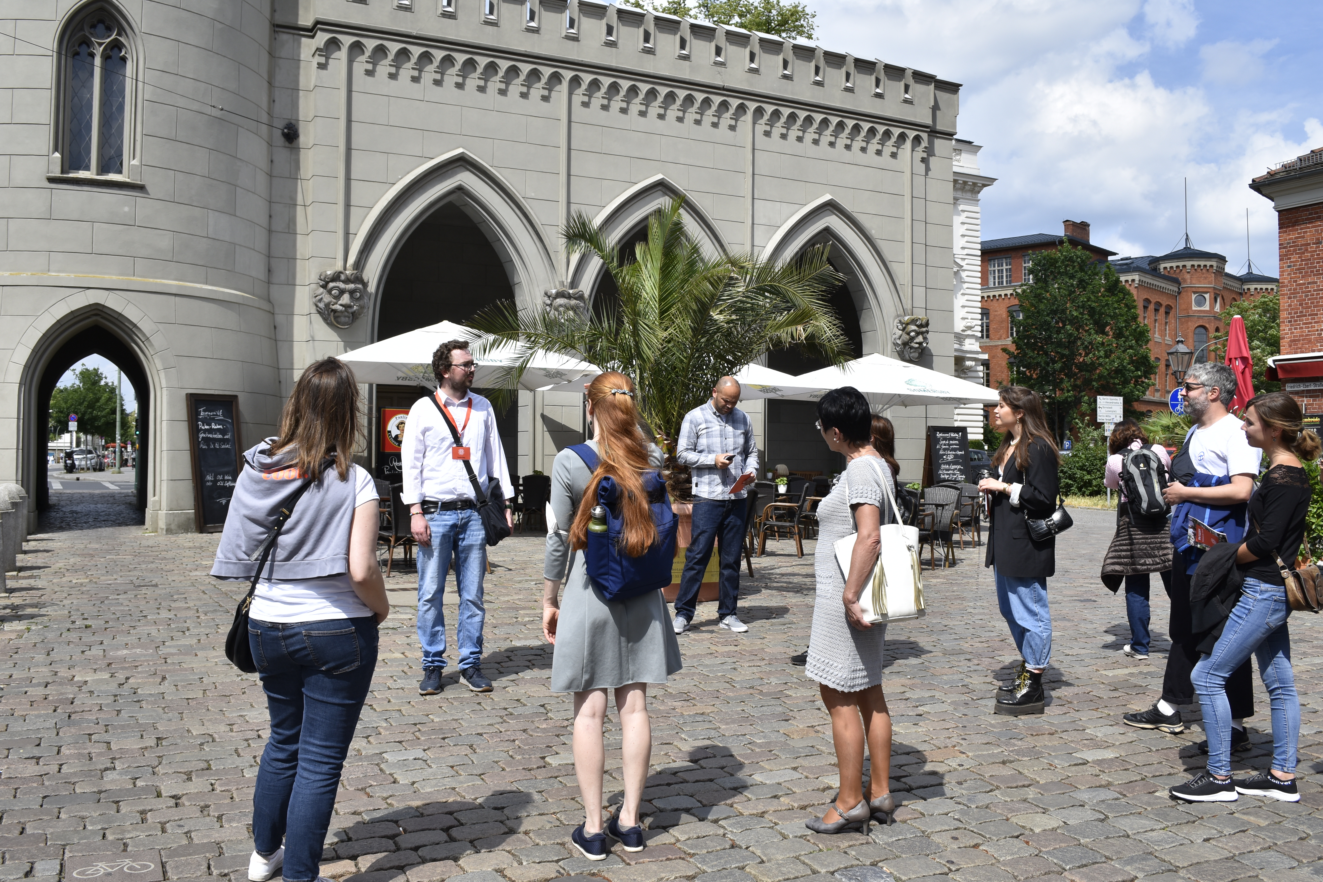 Eine Gruppe von Menschen steht auf einem Kopfsteinpflasterplatz in der Nähe eines historischen Gebäudes mit gewölbten Türen und Sitzgelegenheiten im Freien und genießt ihren Tagesausflug nach Potsdam, während sie die Sehenswürdigkeiten der Stadt erkundet.