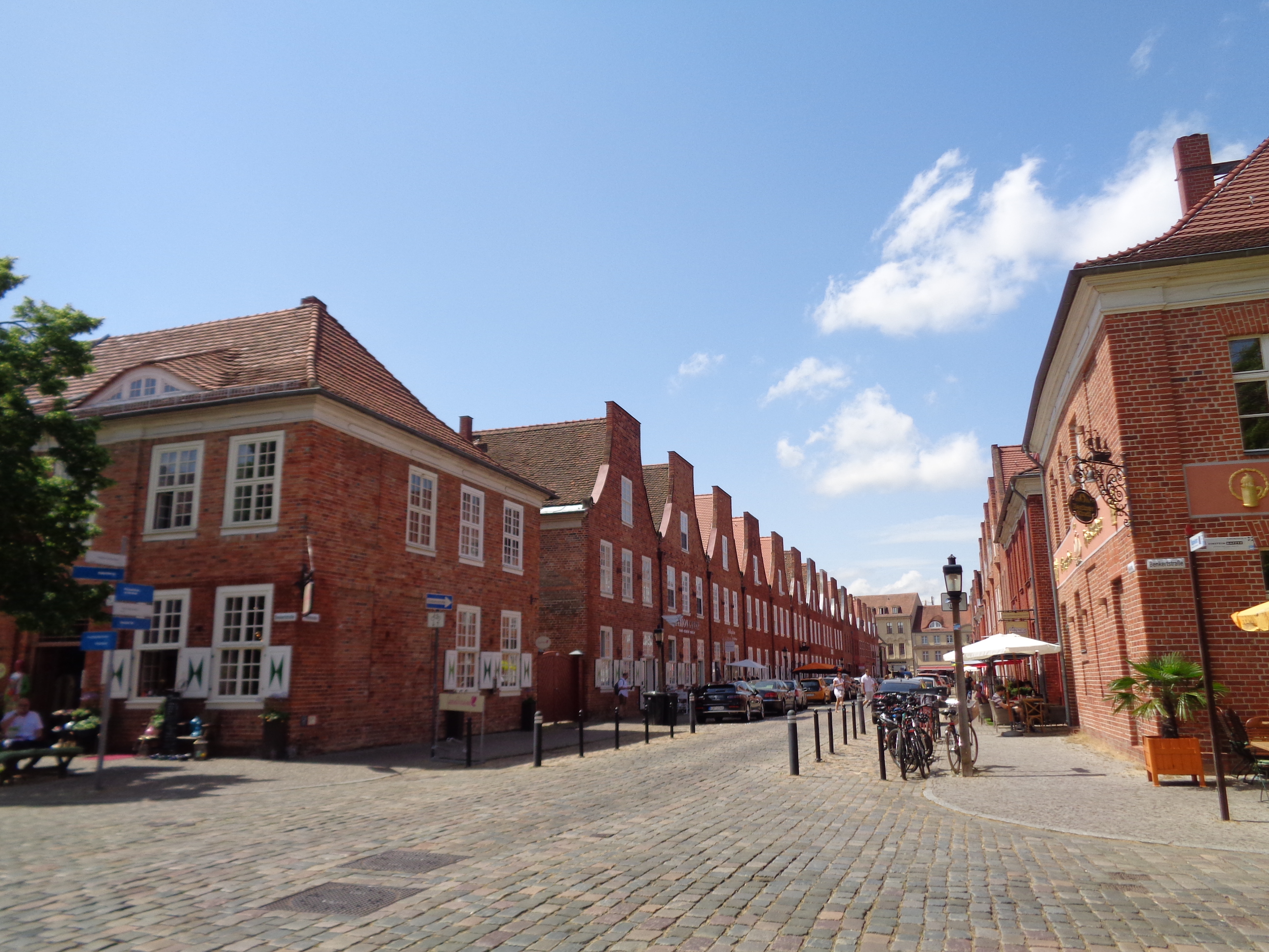 Eine Kopfsteinpflasterstraße in Potsdam, gesäumt von roten Backsteinhäusern und Straßencafés – ideal für einen Tagesausflug unter blauem Himmel mit vereinzelten Wolken.
