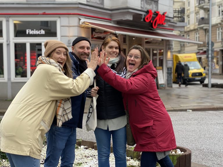 Vier lächelnde Erwachsene in Winterkleidung geben sich draußen auf einer Straße in Köln ein High-Five und feiern eine erfolgreiche Schnitzeljagd.