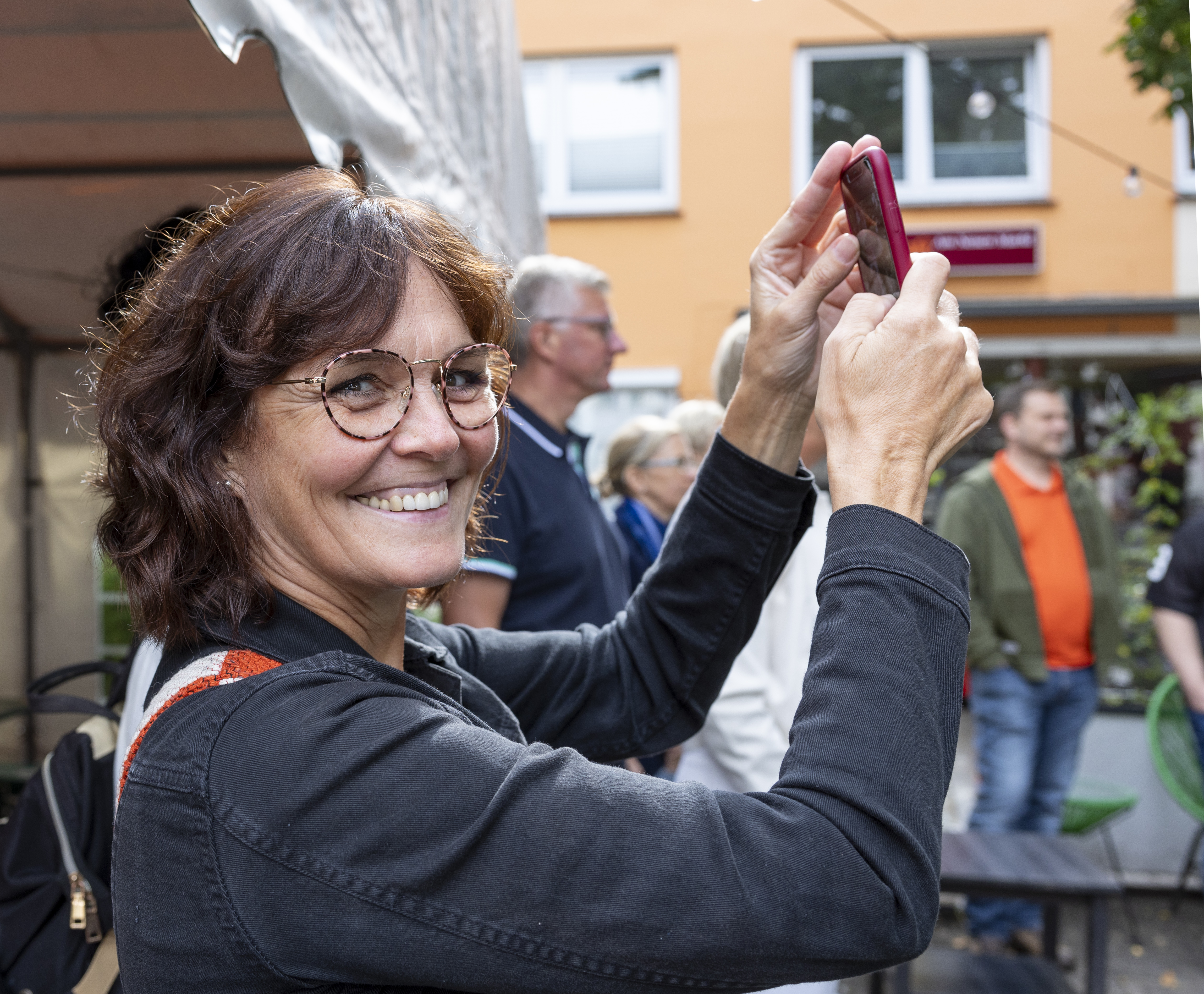 Eine lächelnde Frau, die bei einer Veranstaltung im Freien ihr Handy hochhält, um ein Foto zu machen, im Hintergrund sind Menschen zu sehen – perfekt für die über 50-Jährigen, die gemeinsame Erlebnisse schätzen und diese lieber gemeinsam als allein genießen möchten.