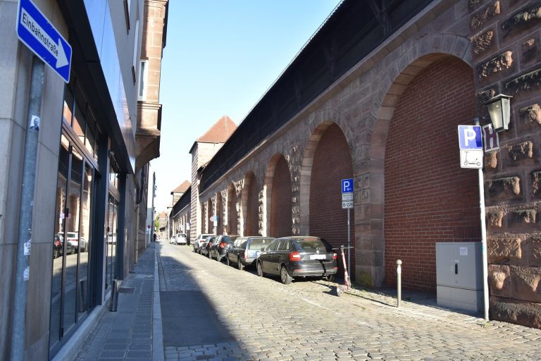 Eine Kopfsteinpflasterstraße mit geparkten Autos neben großen Steinbögen und einem Fahrradabstellschild unter strahlend blauem Himmel, im Herzen der Nürnberger Altstadt nahe der Südlichen Stadtmauer gelegen – ein perfekter Ausgangspunkt für einen historischen Rundgang.