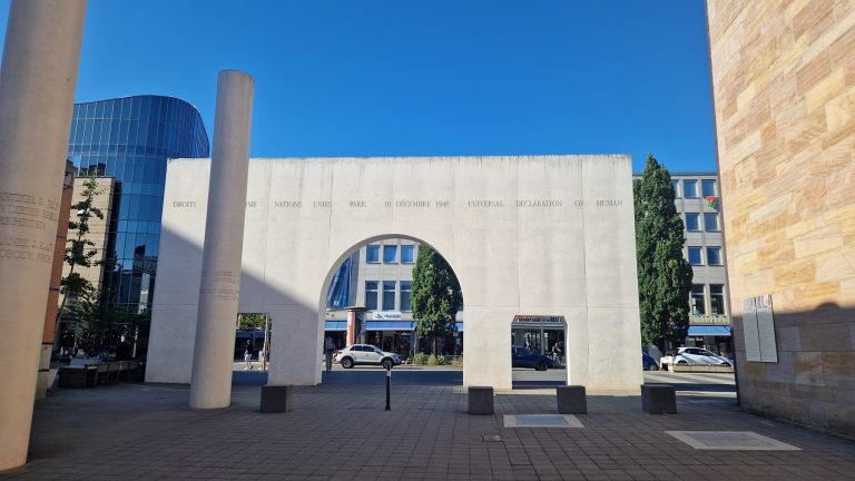 Auf einem Stadtplatz in der Nürnberger Altstadt steht ein Denkmal in Form eines weißen Steinbogens, im Hintergrund sind moderne Gebäude und Bäume zu sehen – es lädt Besucher zu einem historischen Rundgang in der Nähe der südlichen Stadtmauer ein.