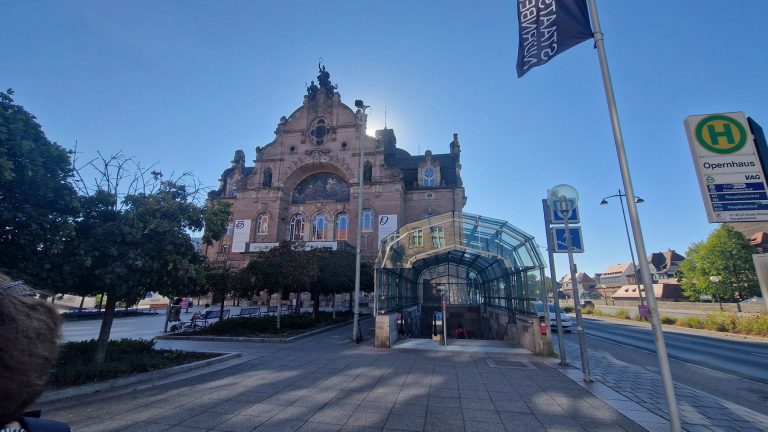 Ein historisches Gebäude mit kunstvoller Fassade hinter einer glasüberdachten Bushaltestelle an einem sonnigen Tag – ein Bild, das den Charme einer historischen Führung durch die Nürnberger Altstadt entlang der südlichen Stadtmauer perfekt einfängt.