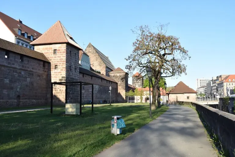 Eine alte Festungsmauer entlang der Südlichen Stadtmauer, ein Baum, ein Weg und eine Grünfläche unter strahlend blauem Himmel an einem sonnigen Tag – ideal, um die Nürnberger Altstadt auf Ihrer historischen Tour zu erkunden.