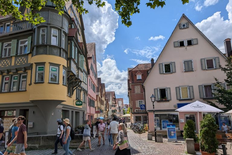 Unter einem strahlenden, teilweise bewölkten Himmel spazieren Menschen auf einer Kopfsteinpflasterstraße, die von bunten Gebäuden gesäumt ist, und genießen einen typischen Genießer-Tag in Münster.
