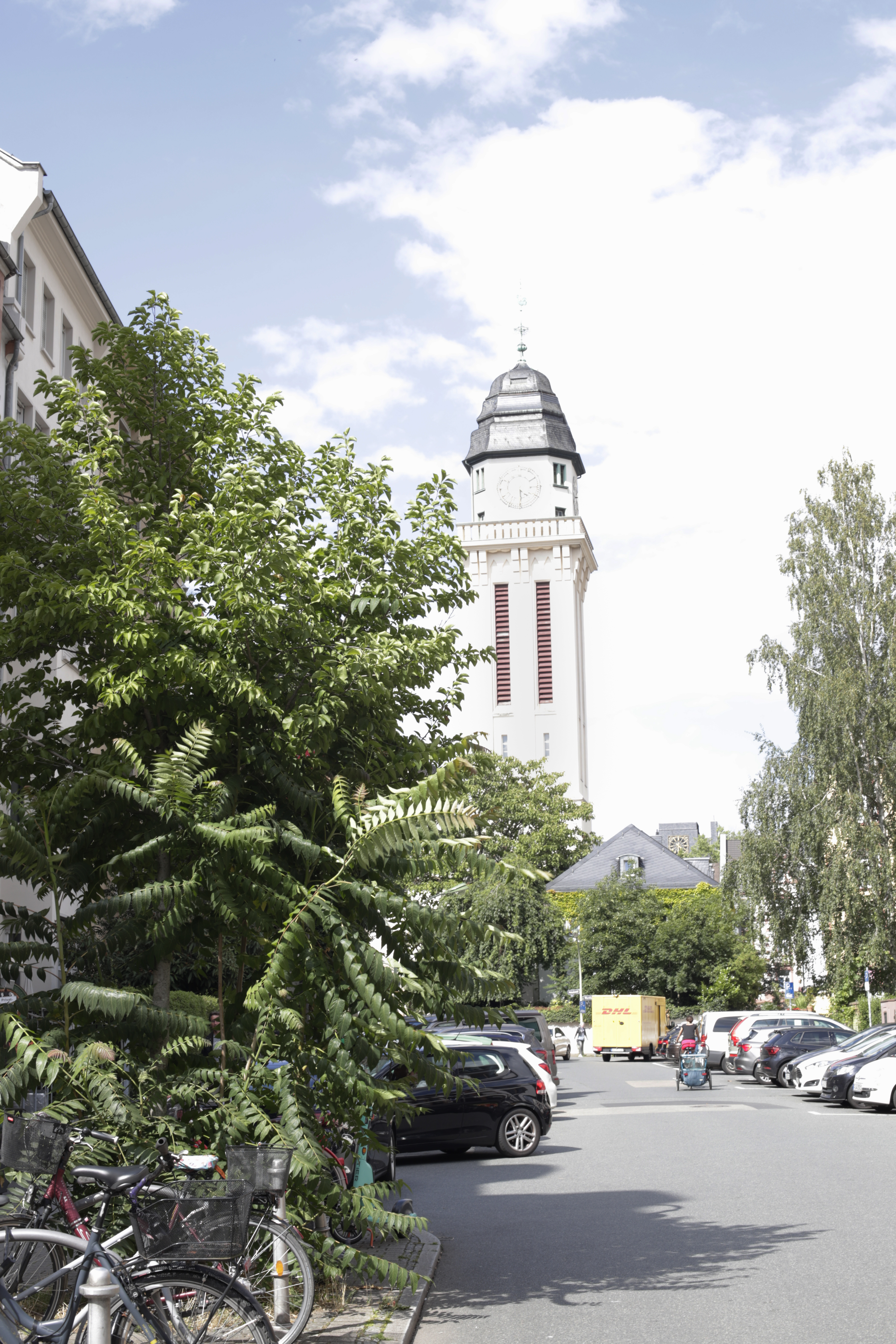 Ein hoher Uhrturm ragt über den Bäumen und geparkten Autos einer sonnigen, teilweise bewölkten Straße empor und bietet Besuchern einen Einblick in „Natur Frankfurt“ – ideal für alle, die „schöne Routen in Frankfurt“ oder entspannte „Spaziergänge in Frankfurt“ suchen.