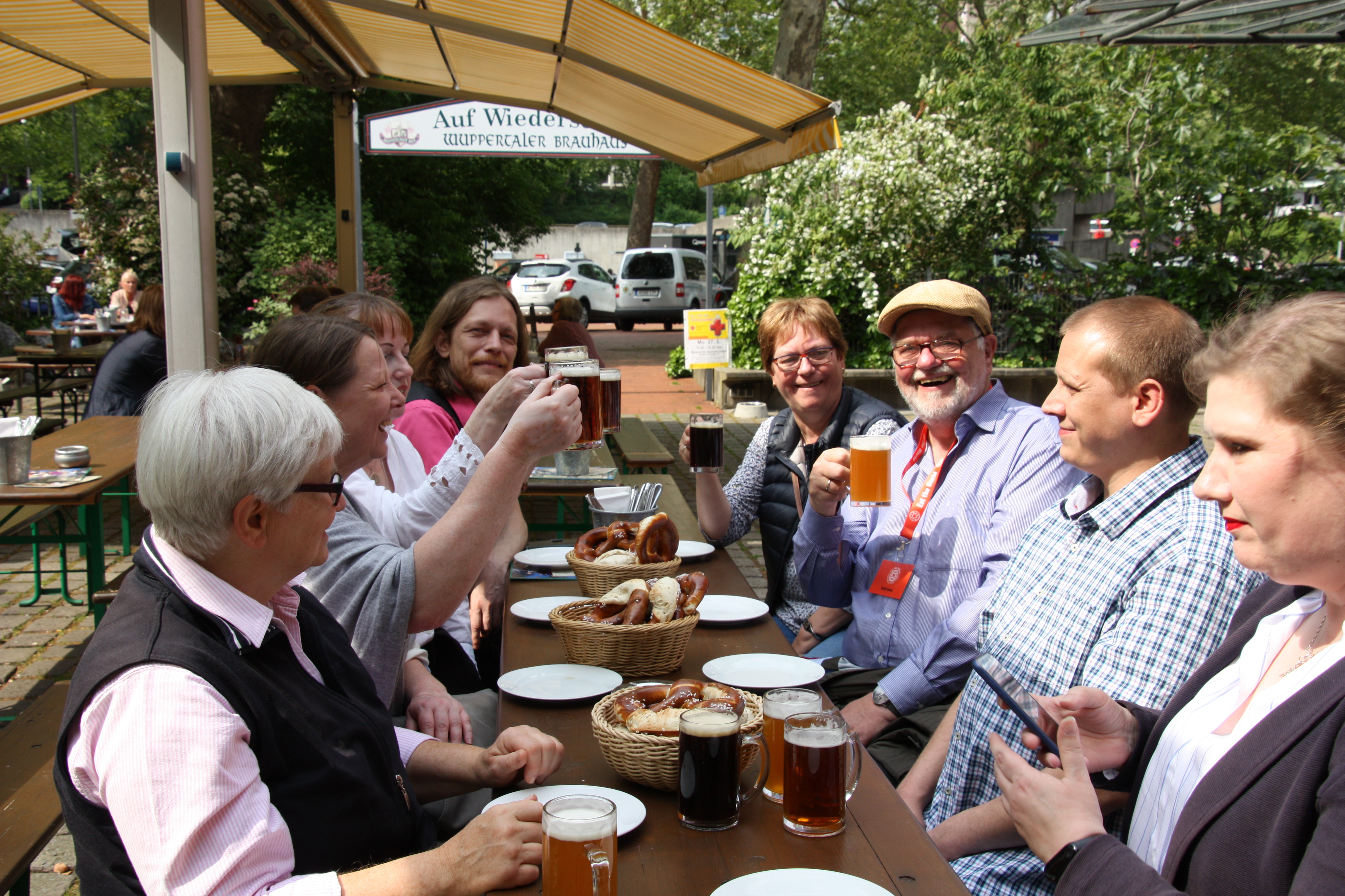 Acht Menschen sitzen an einem Tisch im Freien mitten in der belebten Altstadt von Wuppertal-Elberfeld, stoßen an und lächeln, während Essen und Bier auf dem Tisch stehen.