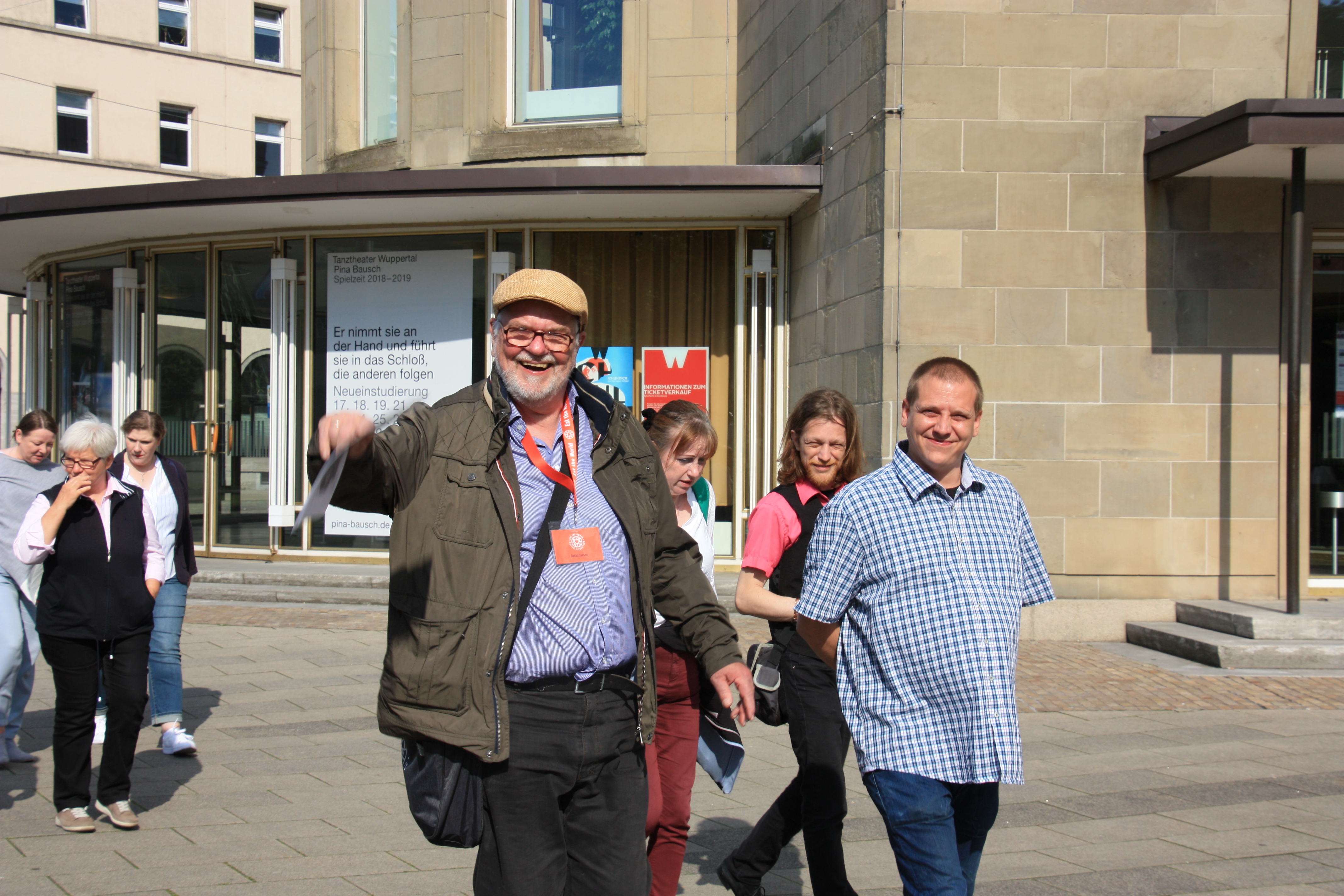 An einem sonnigen Tag in Wuppertal-Elberfeld spaziert eine Gruppe von Menschen durch die Straßen; ein Mann ganz vorne lächelt und winkt, im Hintergrund ist die historische Altstadt gerade noch zu erkennen.