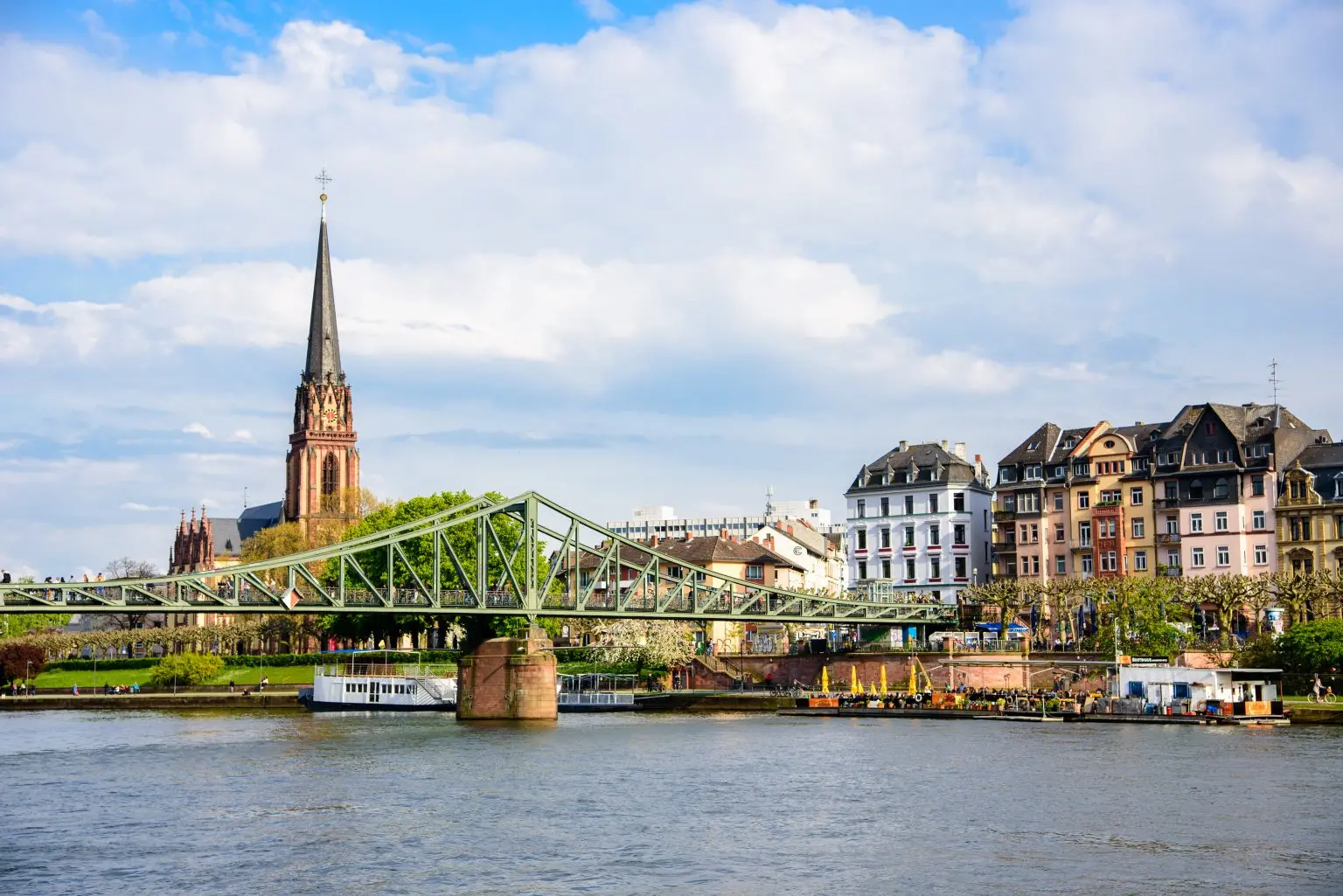 Ein Stadtbild am Flussufer mit einer Brücke, historischen Gebäuden und einem hohen Kirchturm unter blauem Himmel fängt die einzigartige Frankfurter Lebensart ein und hebt die Sehenswürdigkeiten von Frankfurt-Sachsenhausen hervor.