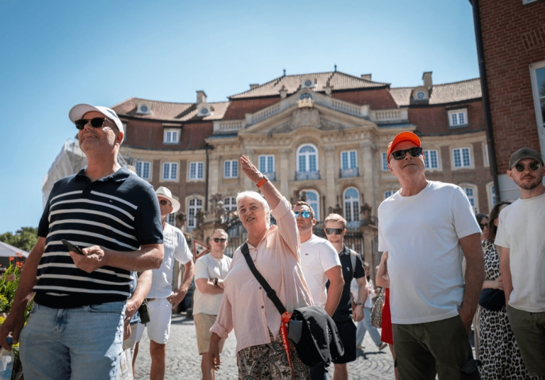 Eine Frau macht Gesten, während sie an einem sonnigen Tag vor einem prunkvollen historischen Gebäude eine Gruppenführung leitet.