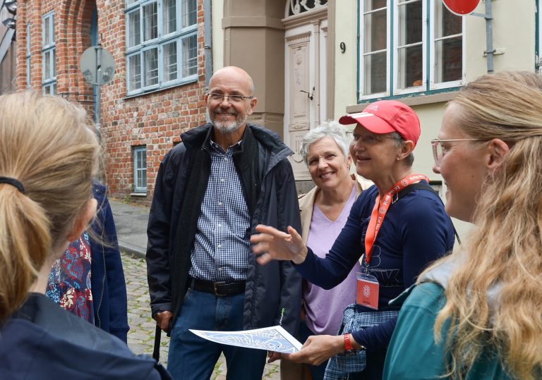 Ein Reiseleiter spricht während einer „Historischen Tour“ im charmanten Lübecker Stadtteil St. Jürgen auf einer Kopfsteinpflasterstraße vor historischen Gebäuden zu einer lächelnden Gruppe von Erwachsenen.
