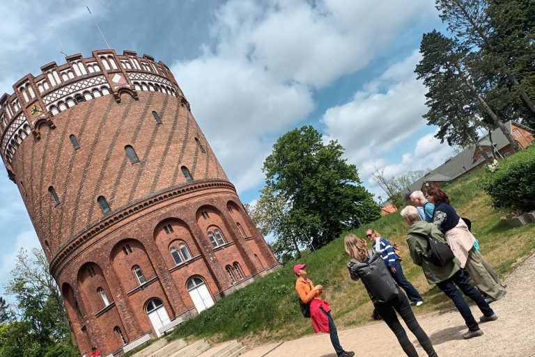 Unter einem bewölkten blauen Himmel lauscht eine Gruppe den Erläuterungen eines Stadtführers in der Nähe eines großen, zylindrischen Backsteingebäudes im Rahmen einer historischen Führung durch den Lübecker Stadtteil St. Jürgen.