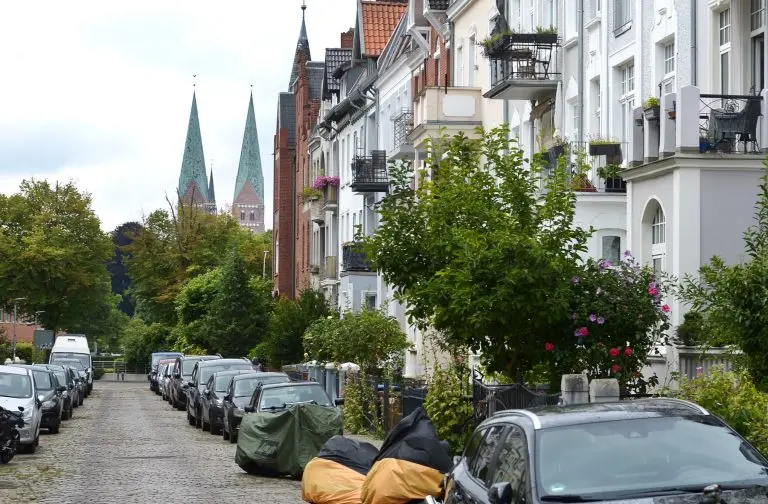 Eine von Bäumen gesäumte Straße im Lübecker Stadtteil St. Jürgen mit geparkten Autos, abgedeckten Motorrädern und im Hintergrund sichtbaren Kirchtürmen – eine einladende Kulisse für eine Historische Tour.